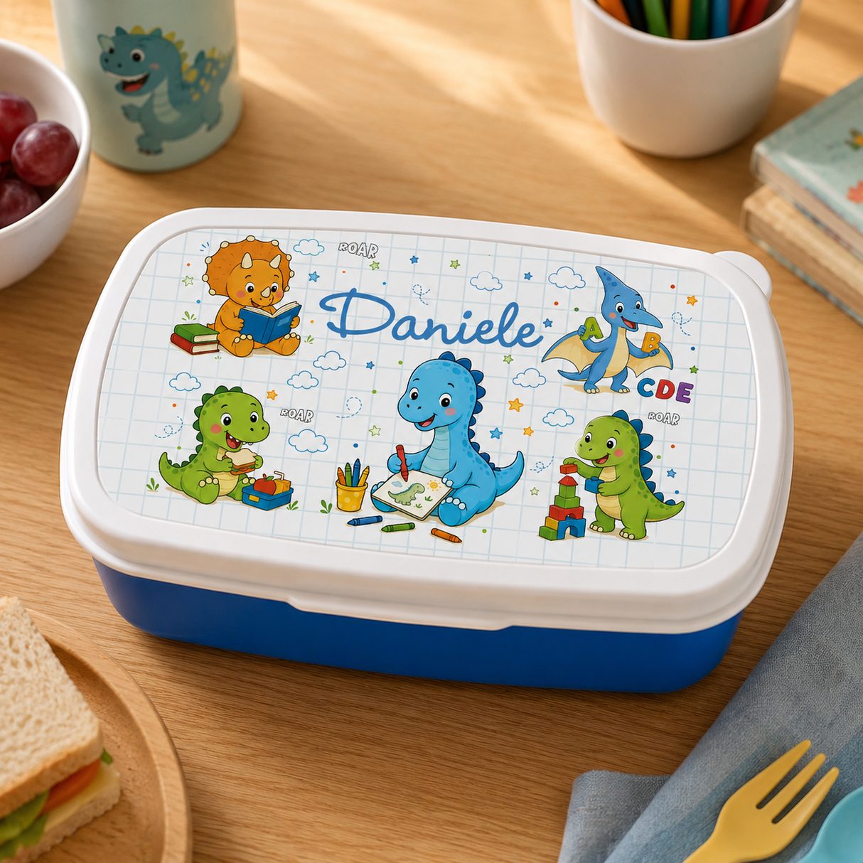 Lunch box blu con coperchio bianco, dinosauri e nome Daniele sul tavolo