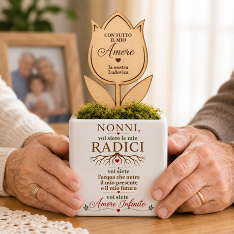 Vaso bianco con fiore in legno inciso tenuto dalle mani di anziani, scritta “Nonni…”.