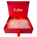 Scatola regalo rossa aperta con scritta “Ti Amo” e riempitivo rosa frantumato